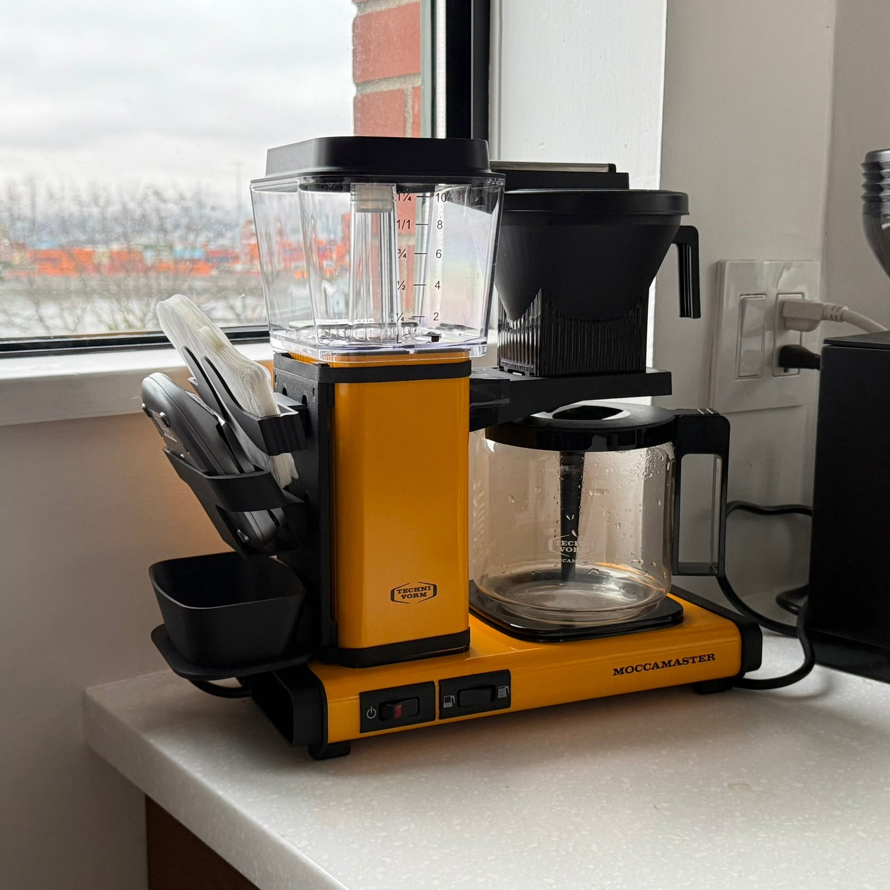 Yellow Moccamaster coffee maker with MoccaBoard modular accessory system holding filters, scale holder, and brewing tools.